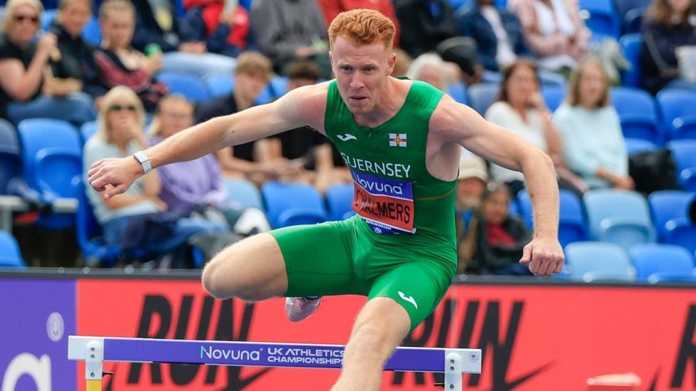 Alastair Chalmers jumps over a hurdle while competing. He is wearing a green Guernsey-branded sleeveless top and shorts